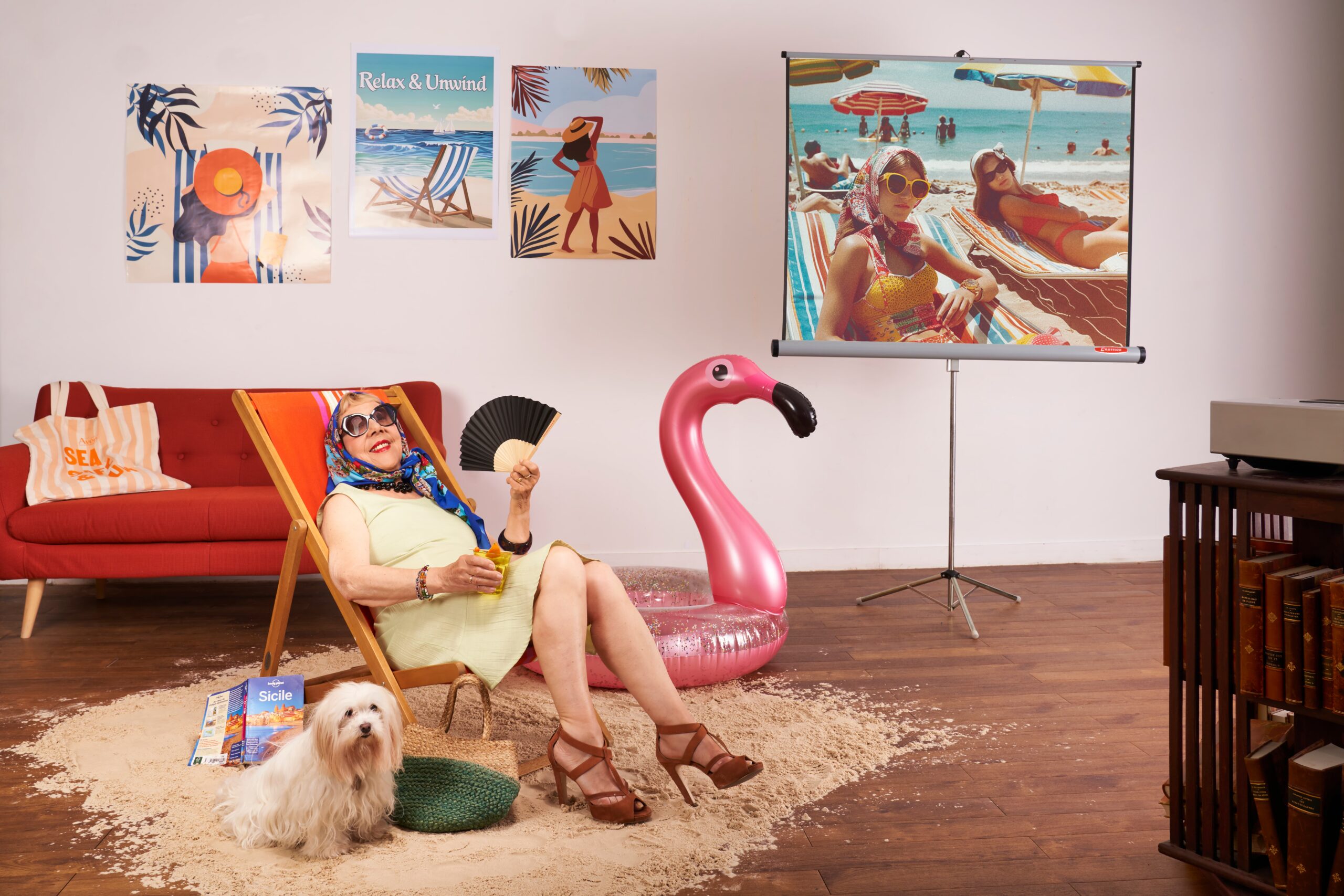 Une femme âgée élégante, coiffée d'un foulard et de lunettes de soleil, installée dans un transat avec un cocktail et un éventail, entourée d'un chien blanc, d'une bouée flamant rose et de posters de plage dans un décor de vacances reconstitué en intérieur.
