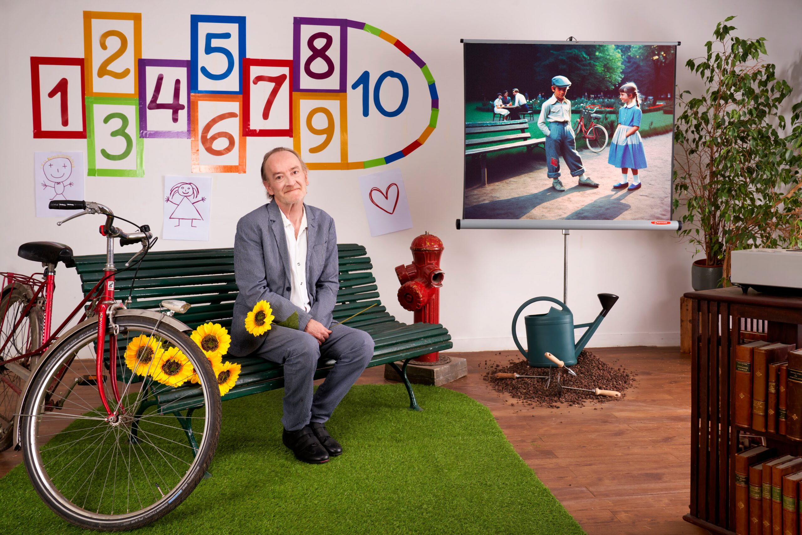 Un homme âgé souriant, assis sur un banc de square vert et tenant un tournesol, dans un décor de jardin public recréé en intérieur avec vélo rouge, marelle colorée, arrosoir et projection d'une scène d'enfants.