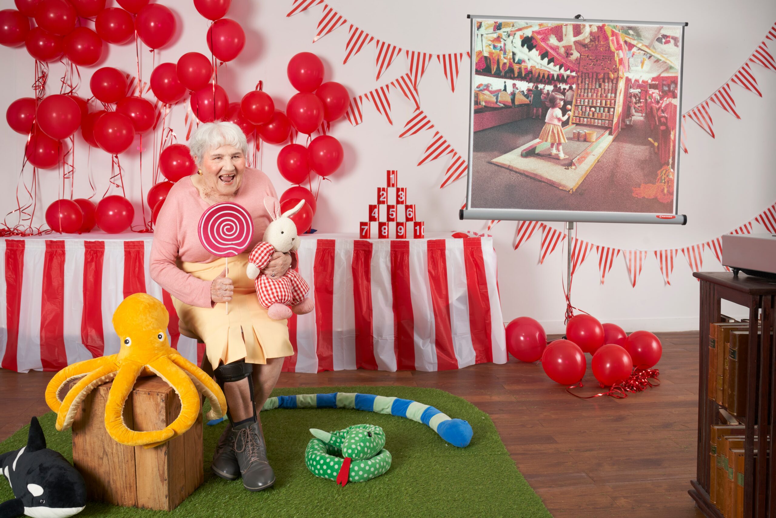 Une femme âgée souriante tenant une sucette géante et un lapin en peluche, entourée de ballons rouges, de fanions rayés et de peluches, dans un décor de stand de fête foraine recréé en intérieur.