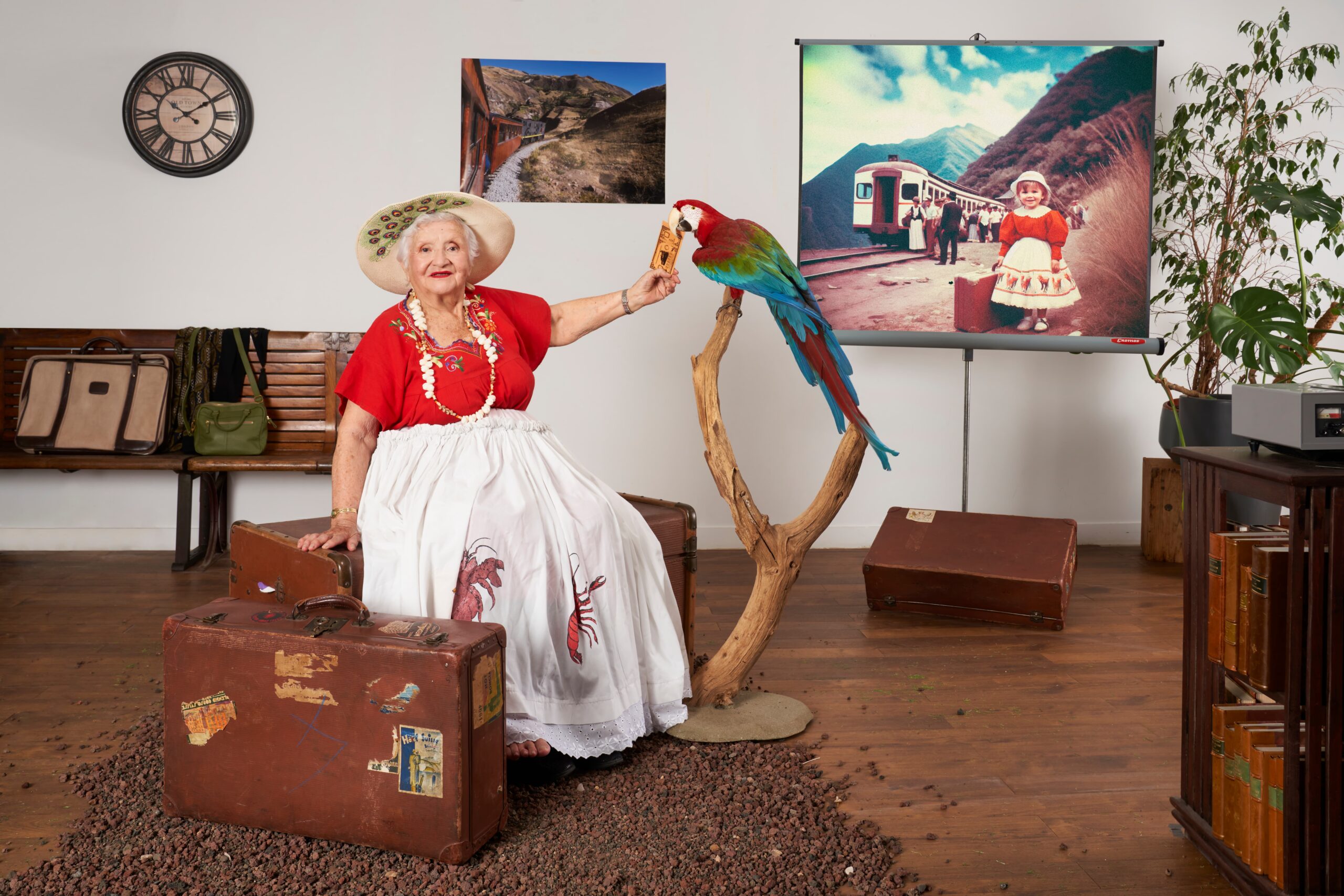 Une femme âgée en tenue folklorique rouge et blanche, coiffée d'un large chapeau, assise sur une malle ancienne, tendant un billet à un perroquet coloré perché à ses côtés, entourée de valises vintage et de photographies évoquant l'Amérique latine.