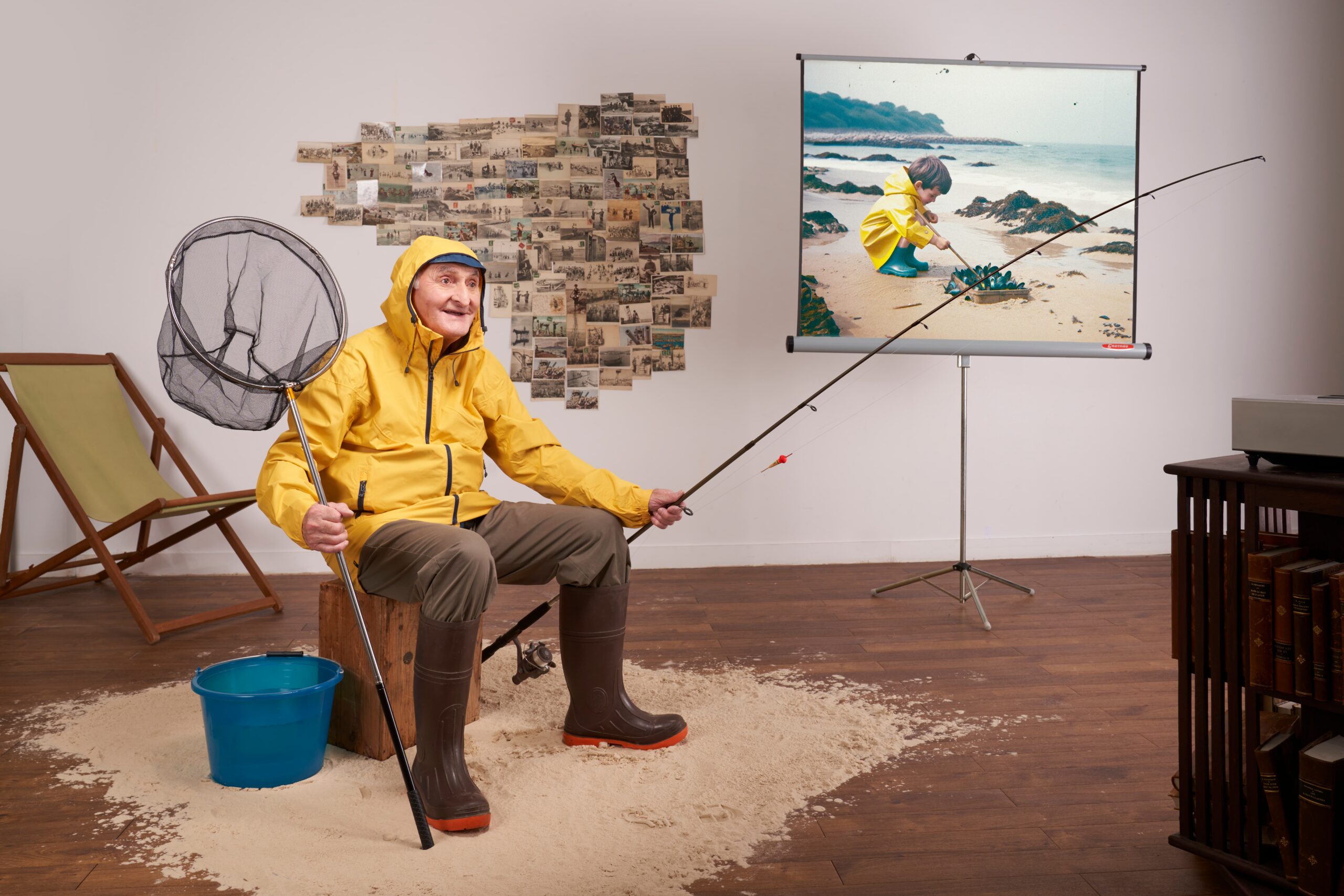 Un homme âgé en ciré jaune et bottes de pêche, tenant une canne à pêche et une épuisette, pose sur un tas de sable dans une pièce, entouré d'une projection d'un enfant au bord de mer et d'une mosaïque de cartes postales anciennes.